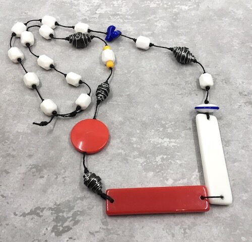 Necklace with red and white long rectangular connectors knotted on black waxed linen with white hex beads, rolled black and grey metal beads, small blue and yellow beads, a red disk and a blue and white disk.